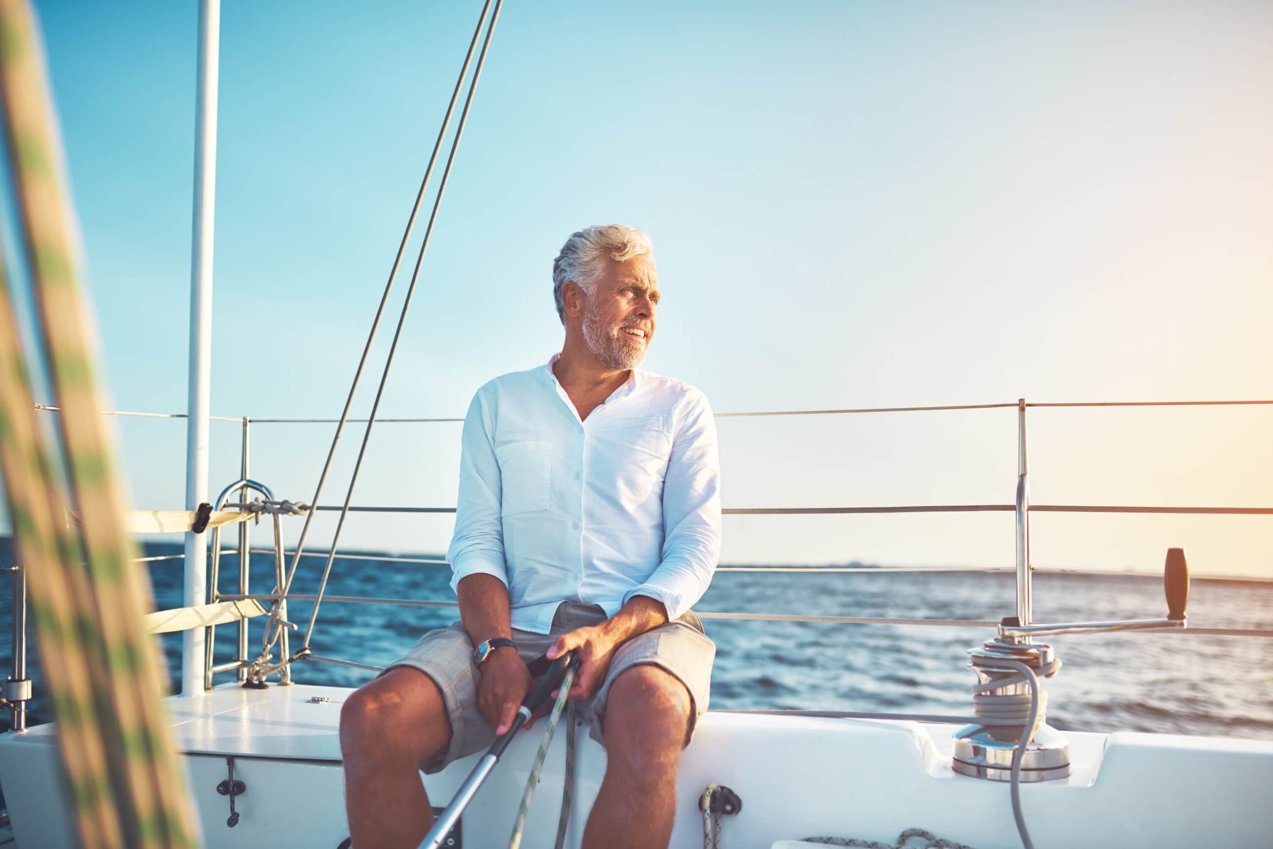 An older man in a white shirt and shorts sitting contentedly on a sailboat, enjoying the view over the ocean under a clear sky. Possibly thinking about selling his boat, and how he could sell with us.