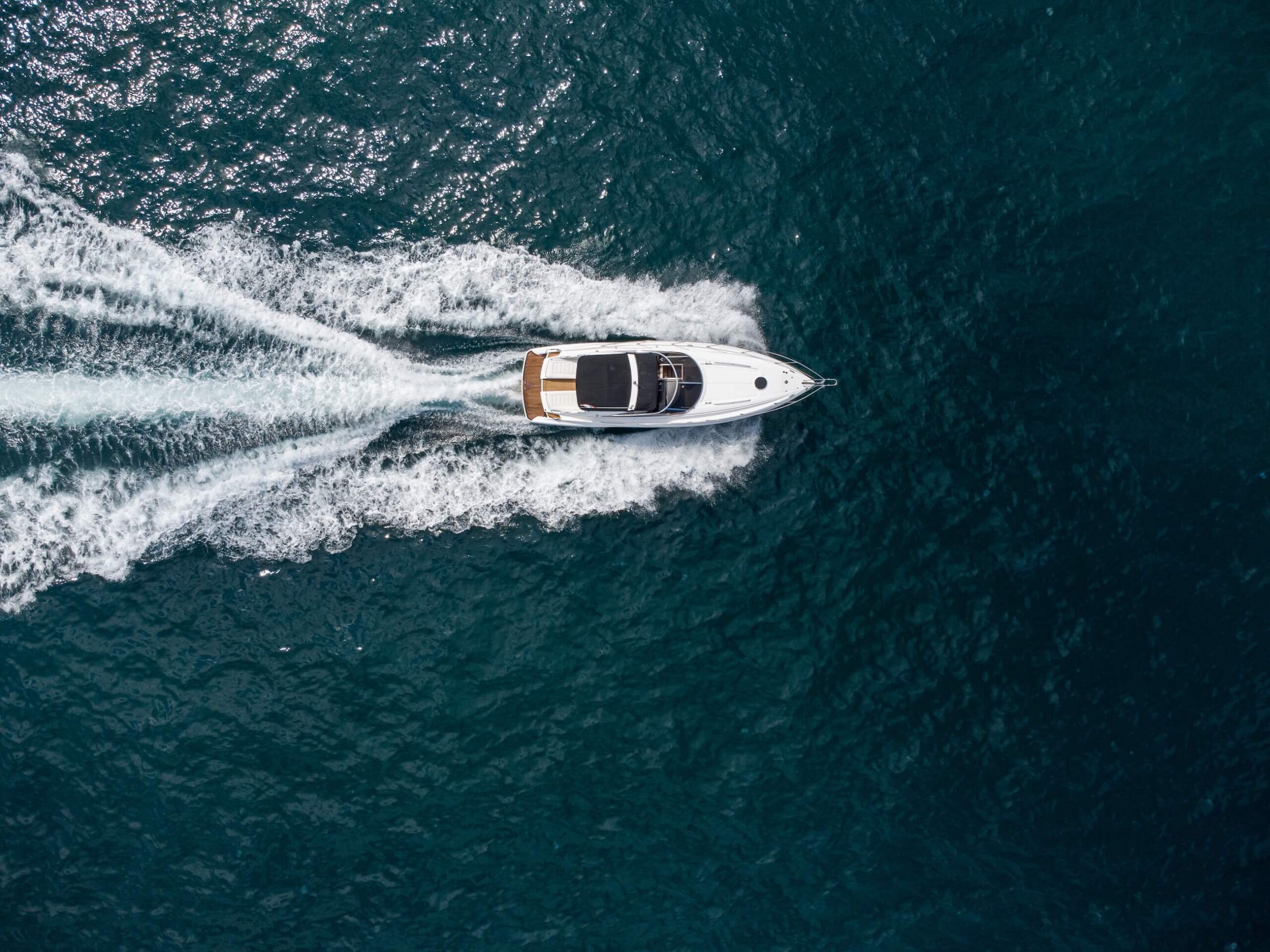 About: Top-down aerial shot of a white speedboat cutting through deep blue water, leaving symmetrical trails of wake behind.